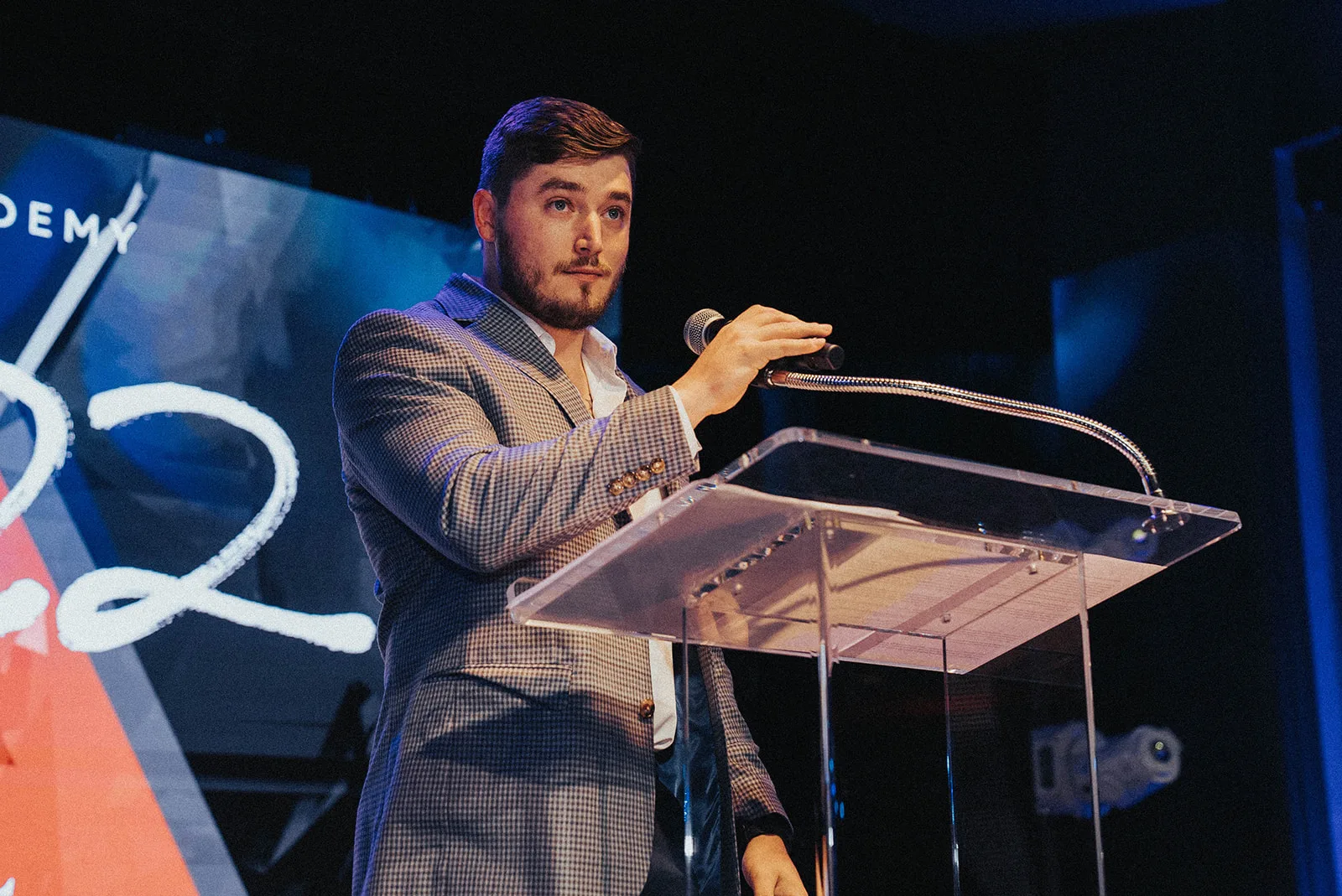 Tyler Dozier on stage at a podium, raising a handheld microphone as he emcees the event
