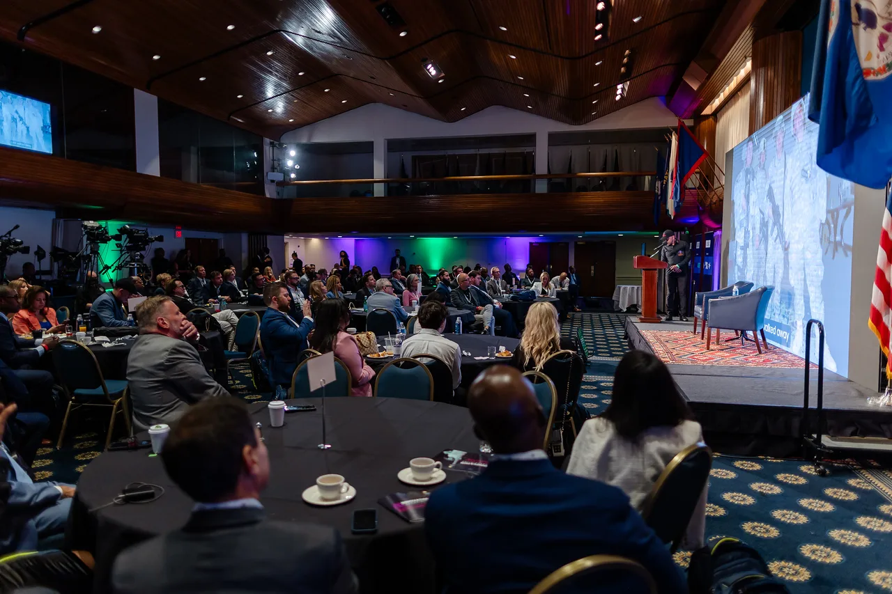 A wide view of a packed general session with a speaker presenting from the stage of a wood-paneled ballroom