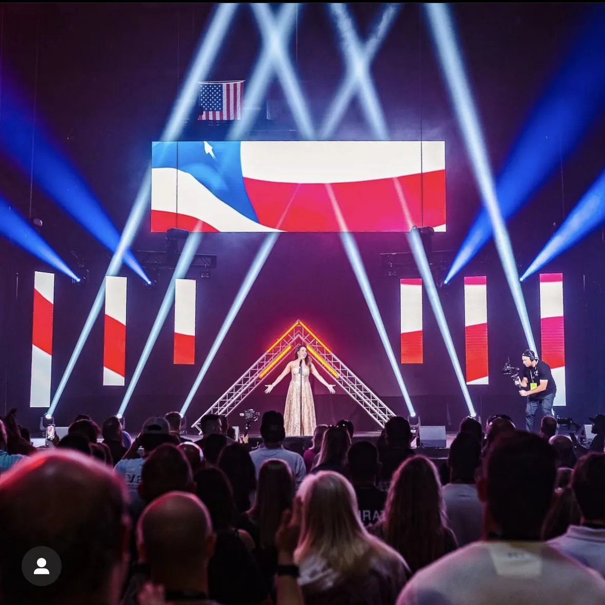 A corporate event stage with a large American flag graphic on screen and white beams of light over the audience