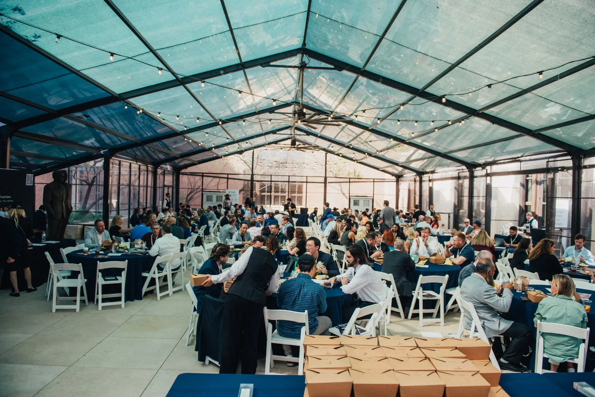 Guests networking over lunch at round tables beneath a glass pavilion during a produced event