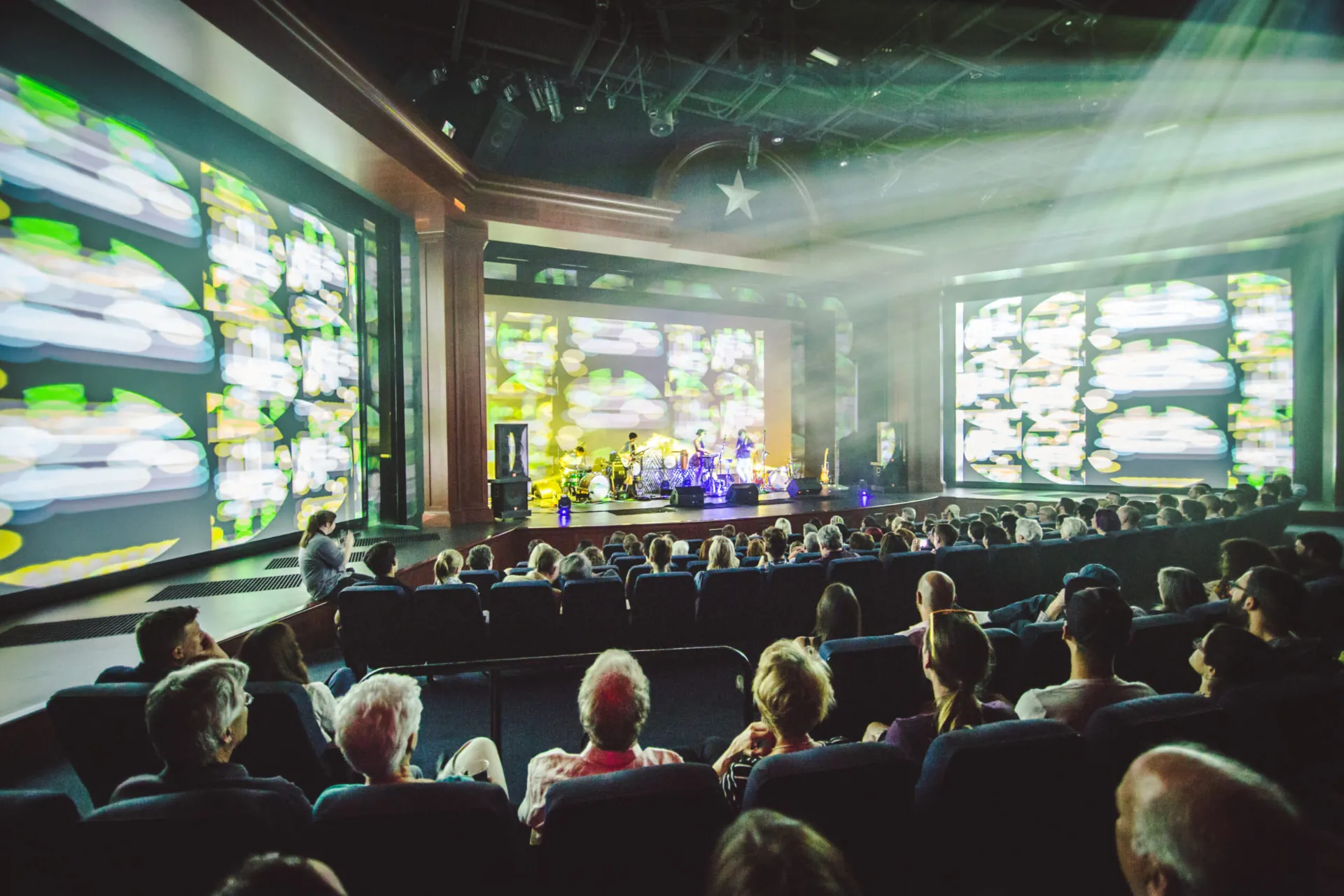 A theater venue with a seated audience facing a stage framed by large video screens